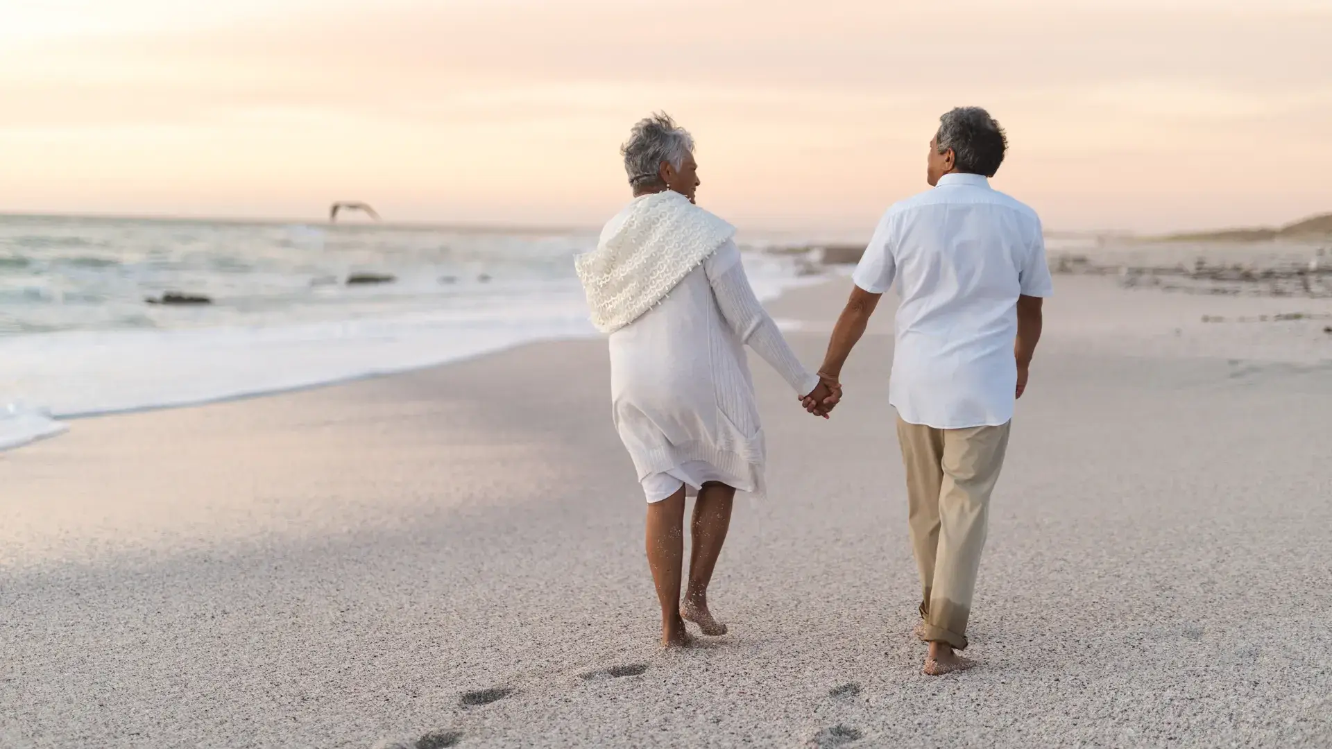Couple de personnes âgées marchant main dans la main sur la plage, symbolisant la paix, la réflexion et le sentiment d’unité.